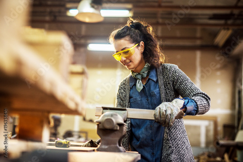 Professional charming cheerful carpenter woman preparing wood on steel vise in the fabric workshop.