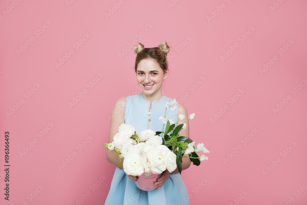 young fashionable woman wearing blue dress holding bunch of flowers, looking at camera while standing on pastel pink color background with copy space. Femininity and beauty