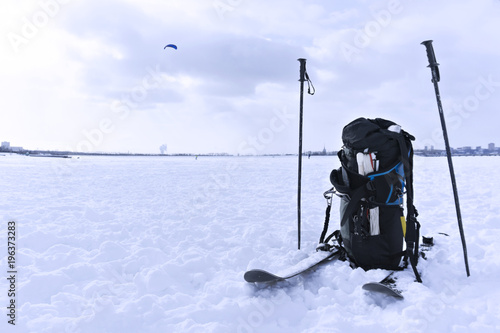 equipment for snowkiting - a backpack with a kite and slings, and skis - on the snow closeup; in the background - gliding snowkiter and city landscape photo