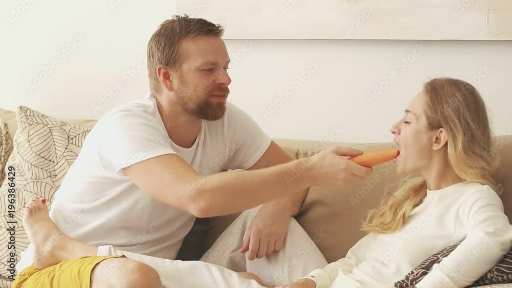 Family couple vegetarian man and woman lying on couch and eating carrot. Family couple vegetarian man and woman lying on couch and eating carrot.