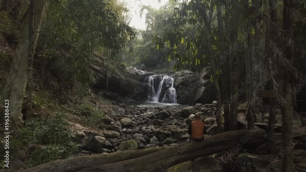 aerial shot man sitting on death tree in front of waterfall in tropical forest at early morning slow camera movement aerial shot man sitting on death tree in front of waterfall in tropical forest at early morning slow camera movement