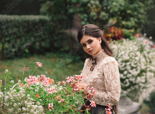 A girl walks in a flowering garden, she has a vintage blouse with a bow, chestnut long hair. she gently cares for her flowers. Sweetheart gardener. Artistic photography. Love of plants