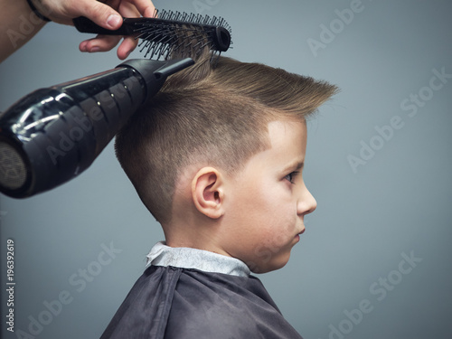 Side view of cute boy getting hairstyle by hairdresser in barbershop.