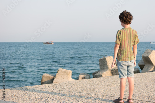The boy is standing on the pier and looking at the sea.