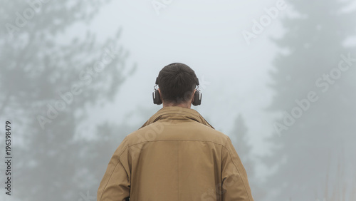 Horizontal rear view of young handsome male walking in misty nature, listening to music outside. Unrecognizable man wearing trendy coat, black bluetooth headphones on head. People, technology concept.