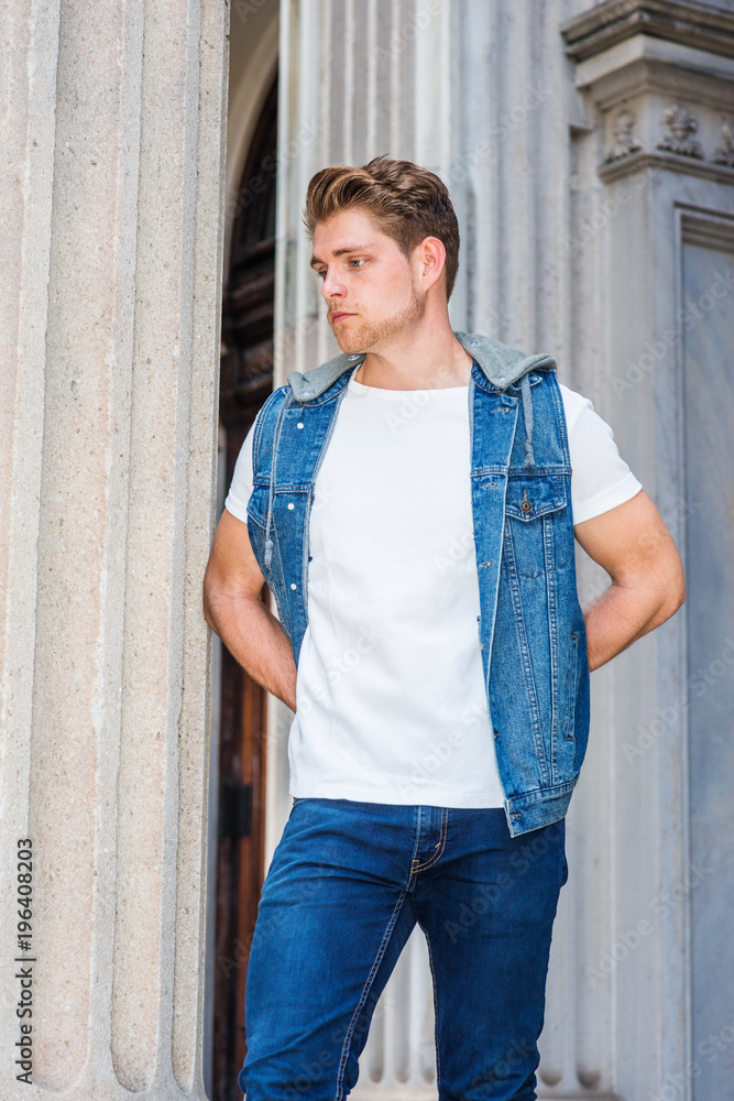 Young American Man with little beard, wearing blue Denim hoody sleeveless  vest Jacket, white T shirt, blue jeans, standing outside vintage office  doorway in New York, hands put in back, thinking.. Stock