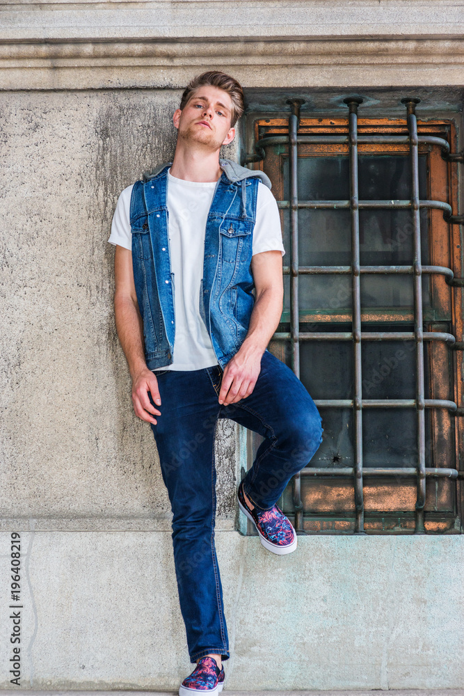 Young American Man with little beard, wearing blue Denim hoody sleeveless vest  jacket, white T shirt, jeans, fashionable shoes, standing against vintage  wall with window in New York. . Stock Photo