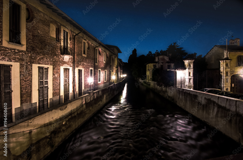 a dark river flows at night between old brick industrial buildings.