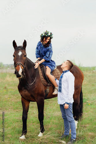 a girl in a beautiful dress and a wreath on her head sits on a horse and looks at the beloved who stands next to her