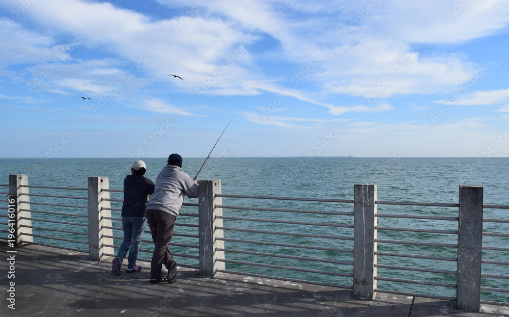 Couple fishing together from pier on sunny day.