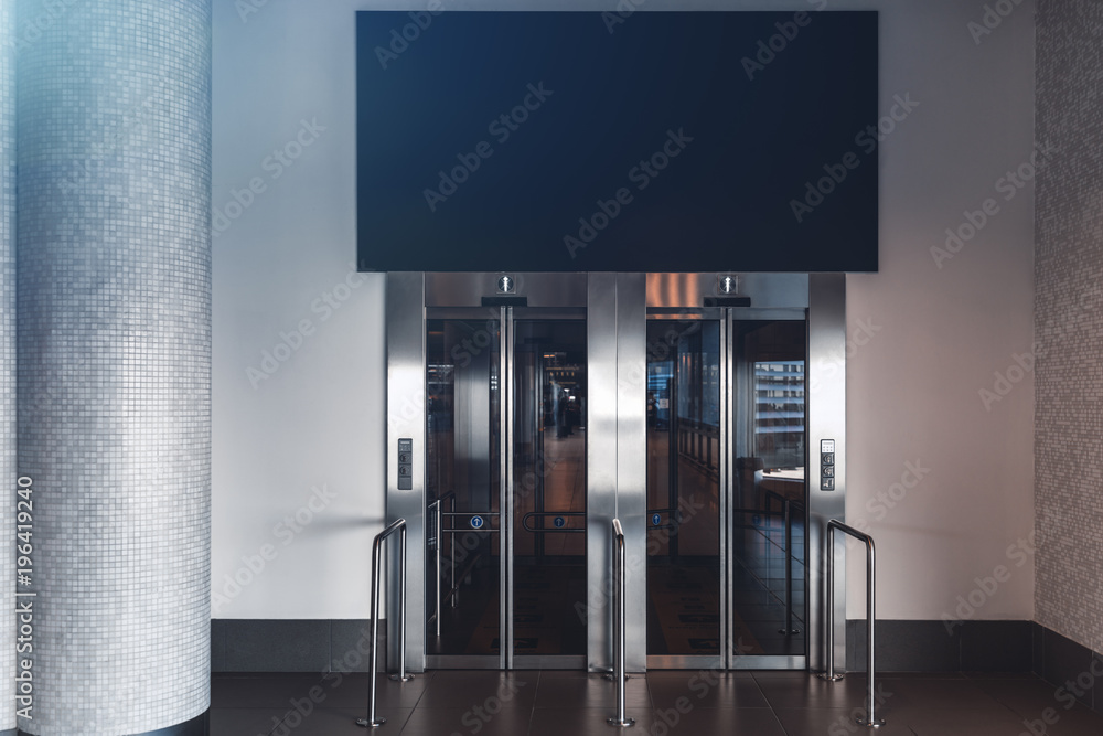 Clean and neat double elevator in a modern airport terminal or a railway depot station with empty blue banner mock-up above; paired lift in a shopping mall with the template of the banner over it Clean and neat double elevator in a modern airport terminal or a railway depot station with empty blue banner mock-up above; paired lift in a shopping mall with the template of the banner over it