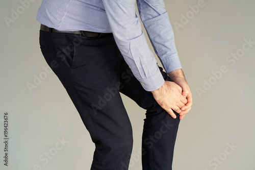 Pain In Knee. Close-up Businessman Leg With Painful Kneeson on gray background. Man Feeling Joint Pain, Having Health Issues And Touching Leg With Hands. Body And Health Care Concept.
