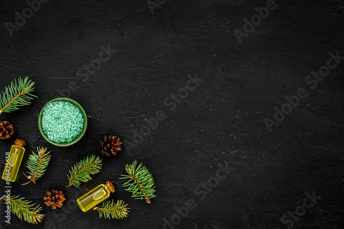 Pine spa cosmetics, products for skin care. Fir essential oil and green aromatic spa salt near branches and cones on black background top view copy space