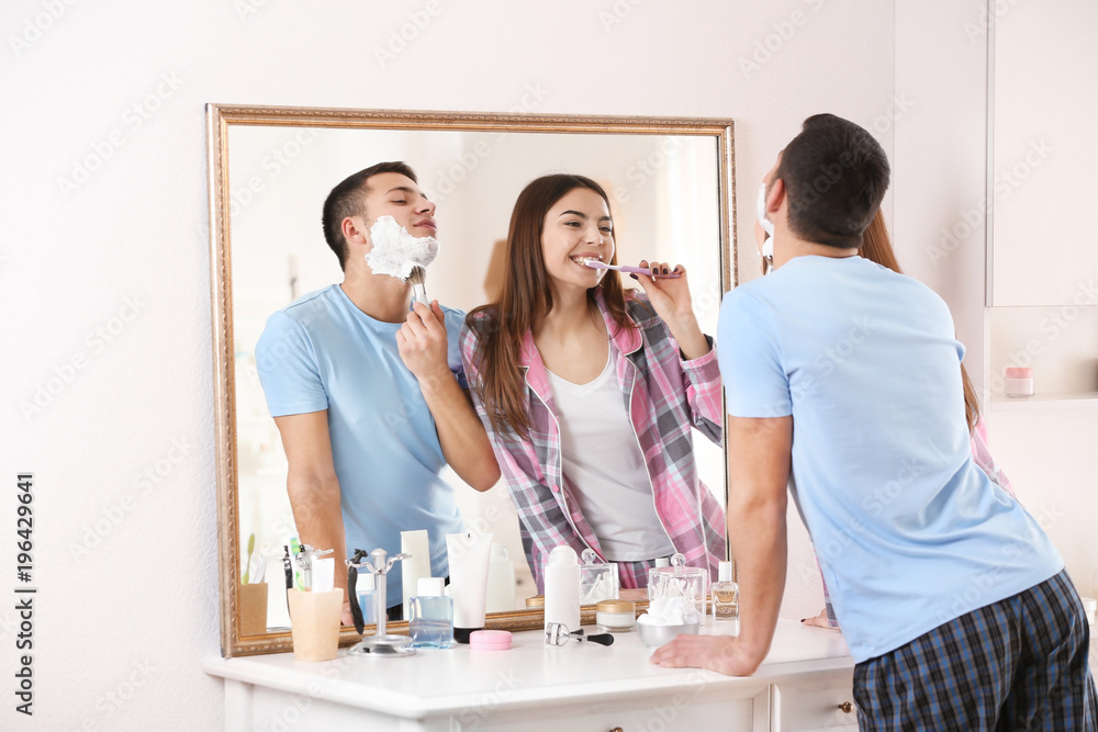 Fototapeta premium Young man shaving and his girlfriend cleaning teeth in bathroom