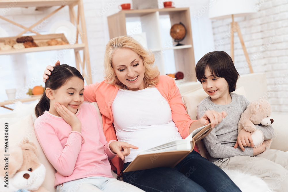 Beautiful grandmother looks at photo album with joyful granddaughter and grandson holding teddy bear.