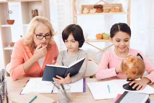 Small beautiful girl is studying globe next to elderly woman who looks at book with little boy.