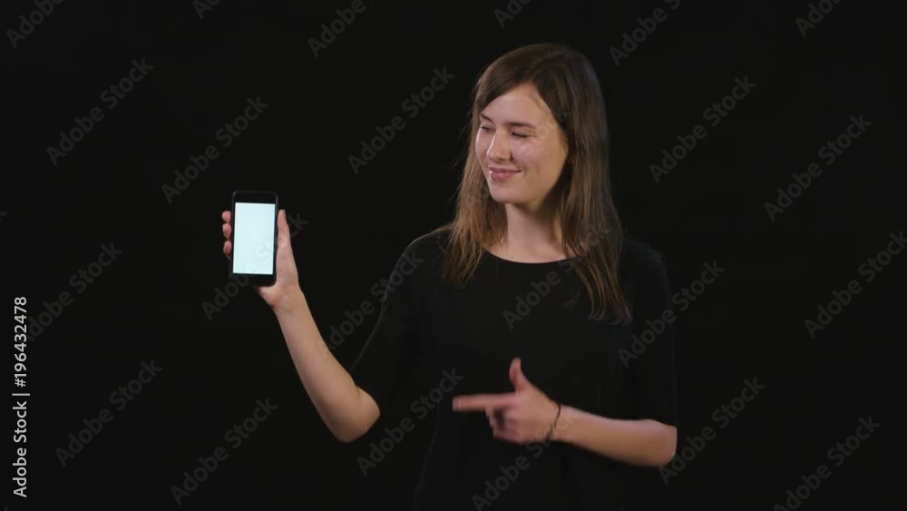 A beautiful young lady pointing her finger at a phone with a white screen against a black background. Medium Shot A beautiful young lady pointing her finger at a phone with a white screen against a black background. Medium Shot