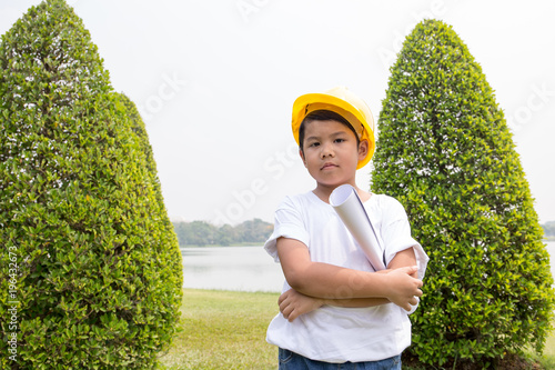 Asian boy in white t-shirt with yellow helmet dress like engineer cross arms to his chest with garden scene