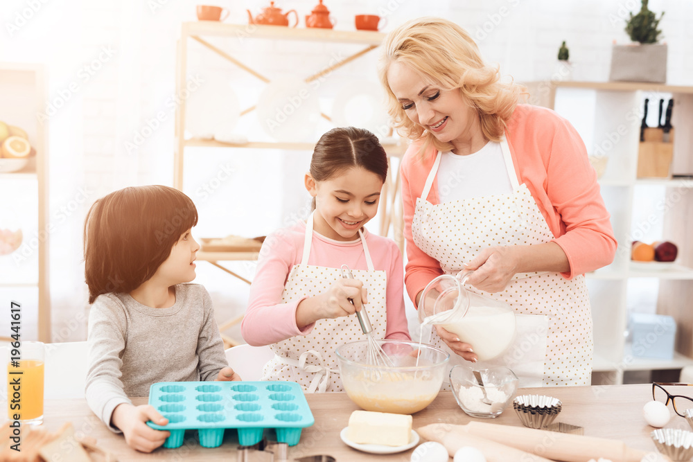 Small cheerful girl is beating eggs in kitchen. Grandmother pours milk into bowl. Small cheerful girl is beating eggs in kitchen. Grandmother pours milk into bowl.