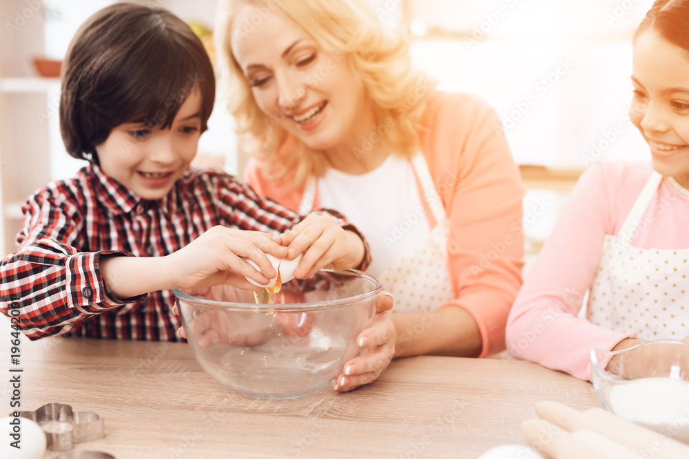 Grandson breaks egg into bowl, sitting next to grandmother and sister sitting behind laptop. Grandson breaks egg into bowl, sitting next to grandmother and sister sitting behind laptop.