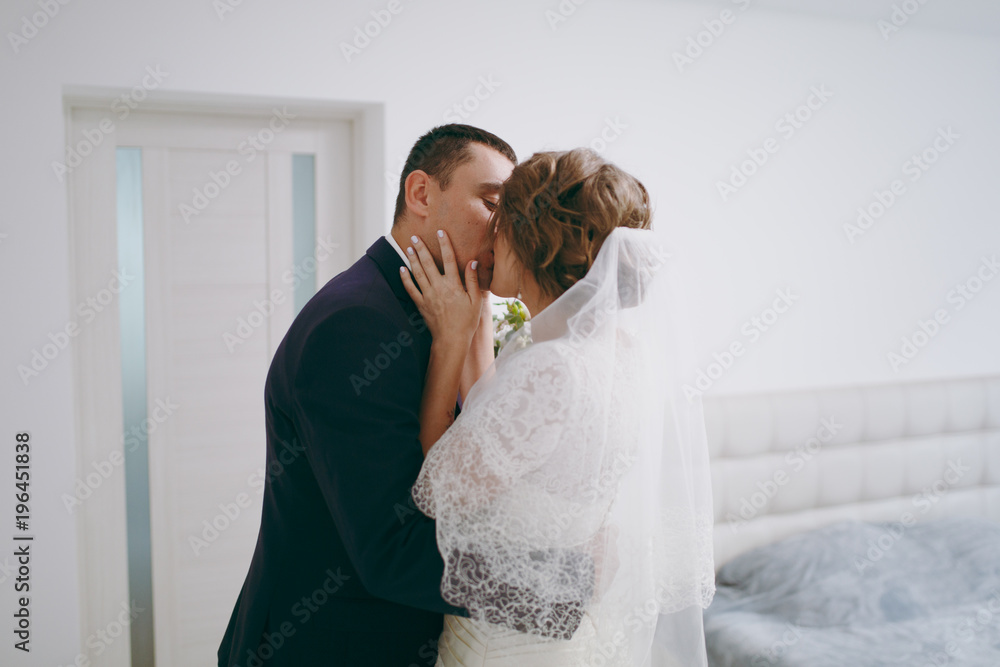 Morning preparation of the newlyweds for the wedding ceremony. Meeting the bride and groom The fiance in a formal suit kisses his fiancee in a white dress and veil with beautiful hairdress in room