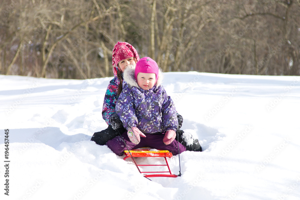 Two kid sisters sitting together on a sled in a snowdrift on a clear winter day. Kids playing outdoors in winter Two kid sisters sitting together on a sled in a snowdrift on a clear winter day. Kids playing outdoors in winter
