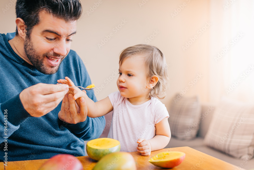 Father feeding toddler baby infant with spoon and mango