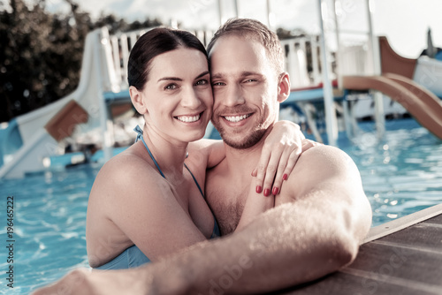 Love is in the air. Beautiful young woman hugging her boyfriend tightly while both standing in a swimming pool and grinning broadly into the camera. © zinkevych