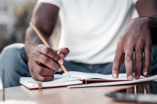 Preparation process. Scaled up look on hands of a young man holding a pencil and writing down some necessary information while sitting outdoors. photo