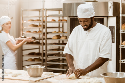 handsome african american baker kneading dough while his female colleague working on background at baking manufacture