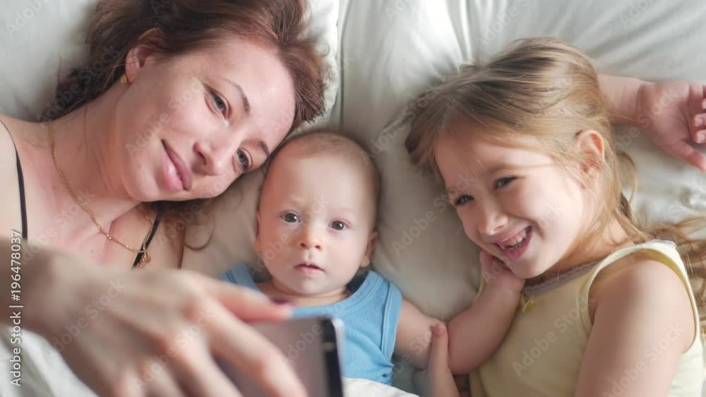 Happy mother and her young children taking selfie with smartphone on bed Happy mother and her young children taking selfie with smartphone on bed