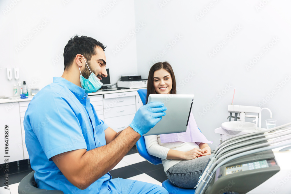 Bright and handsome doctor showing teeth x-ray to the patient on tablet, tall man in blue scrubs explaining the problem using smart technology, pretty woman in white top sitting in dental chair at the Bright and handsome doctor showing teeth x-ray to the patient on tablet, tall man in blue scrubs explaining the problem using smart technology, pretty woman in white top sitting in dental chair at the