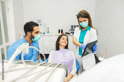 Smiling beautiful patient sitting in chair with dental purple bib  male dentist  in blue uniform and gloves  female assistant  in green mask with tablet  standing next to the patient