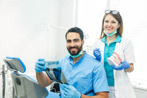 Two dentists discussing particular case, handsome bearded man pointing at teeth x-ray, slim fair-haired girl holding a sample of dentures next to him, indoor shot in modern dentist office