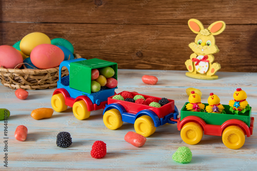 Easter toy car with trailer and color sweets in, Easter eggs, candy and bunny on wooden background Easter toy car with trailer and color sweets in, Easter eggs, candy and bunny on wooden background