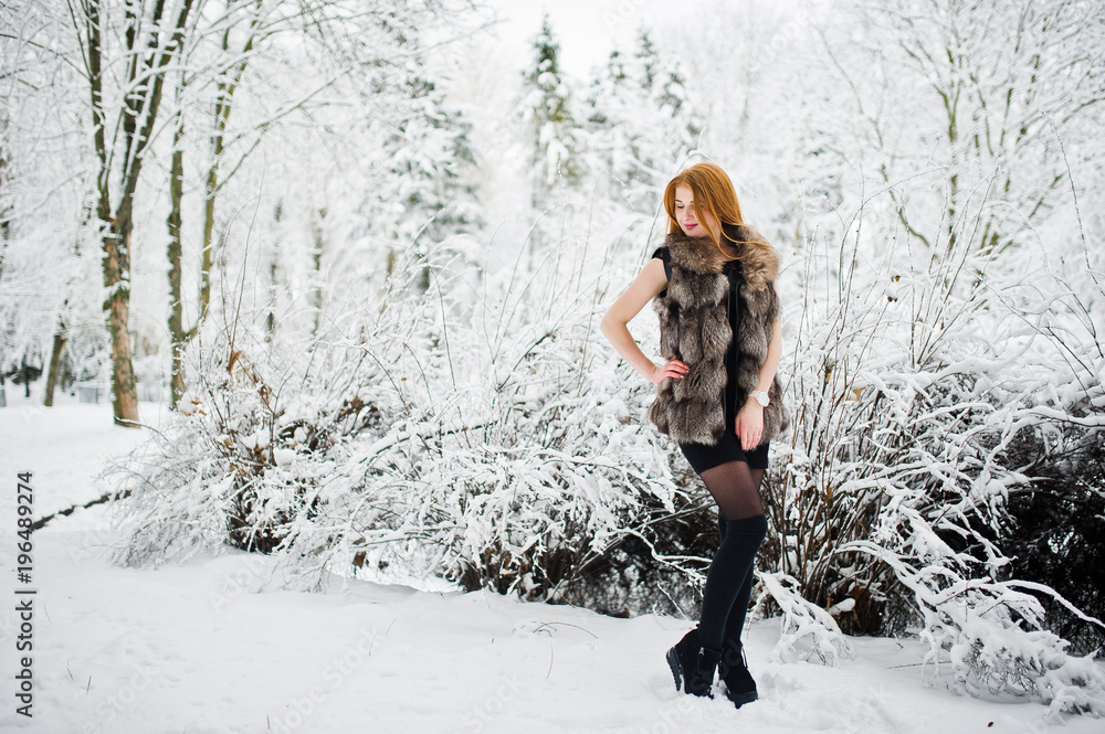 Red haired girl in fur coat walking at winter snowy park.