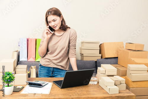 Young happy asian online business woman in her home talking to her customer on her phone while looking at boxes of product