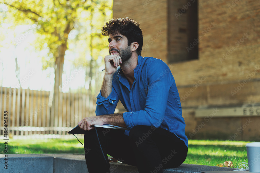 International student preparing for the exams while sitting in a park. Thoughtful bearded man in a light blue shirt sitting with a pencil and a notebook in his hands with a coffee standing beside. International student preparing for the exams while sitting in a park. Thoughtful bearded man in a light blue shirt sitting with a pencil and a notebook in his hands with a coffee standing beside.