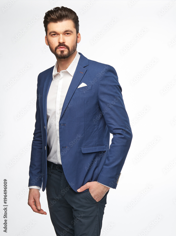 Fototapeta premium Fashion portrait of handsome male model with dark hair, beard and eyes, wearing white shirt, blue jacket and dark pants, posing on white background