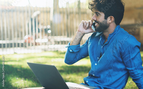 Handsome bearded guy in a laight blue shirt talking by a mobile phone while sitting outdoors with a laptop on his knees. Successful businessman receiving good news about his project by a phone call. photo