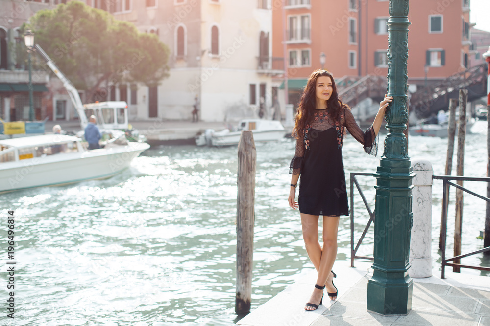 Attractive young romantic woman standing on the pier against beautiful view on venetian chanal with boats and gondolas in Venice, Italy. Travel tourist girl on vacation walking happy by Grand Canal
