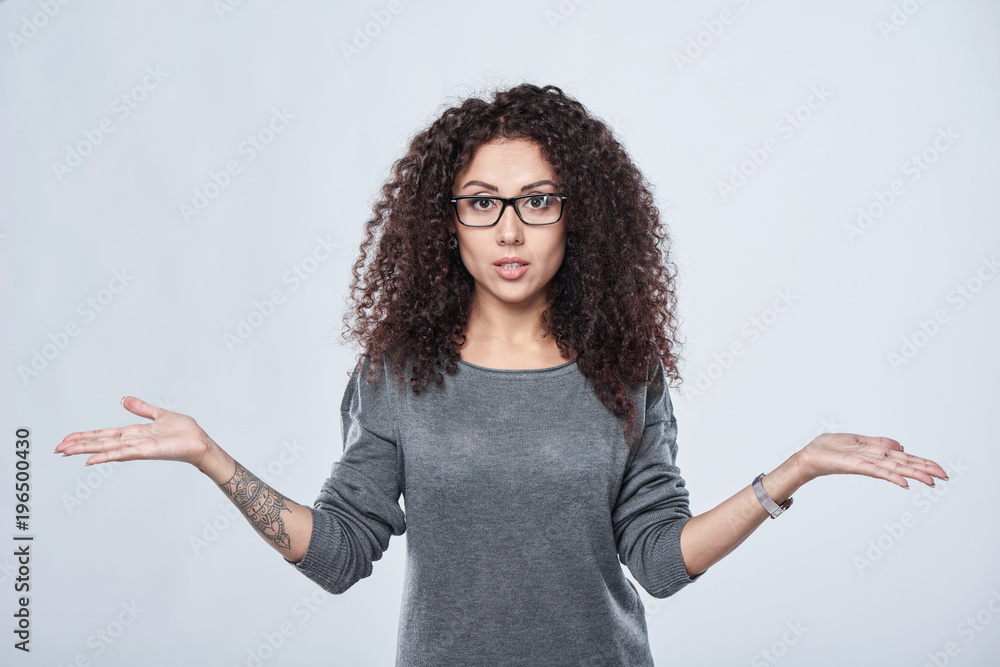 Closeup of confused curly woman in eyeglasses with open spread hands , over grey background Closeup of confused curly woman in eyeglasses with open spread hands , over grey background