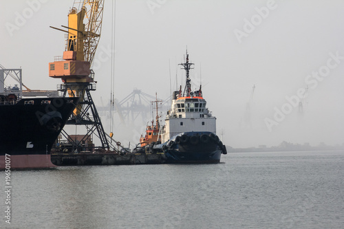  The tug is at the pier in the seaport. The background cranes shrouded in fog