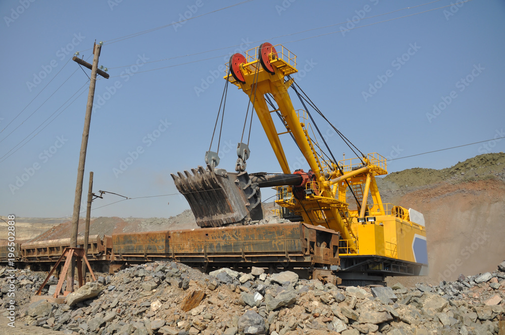A large yellow excavator loads ore into a railway car