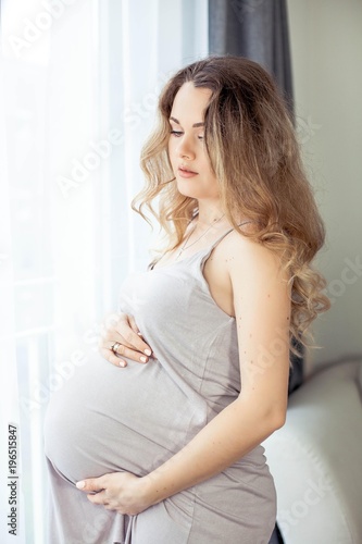 A young beautiful pregnant woman is standing by the window. Pregnant in a beautiful dress. Motherhood. Pregnancy. Portrait of a happy pregnant woman. Cozy.