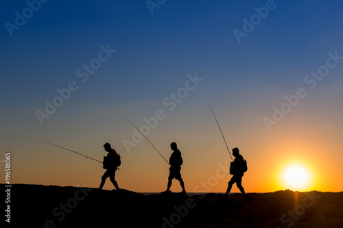 3 Fishermen silhouette with upright rods stroll walk on the rocks at dawn dusk sunrise sunset. The sky is blue purple orange. They have upright rodsat dawn 