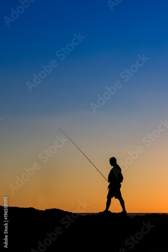 3 Fishermen silhouette with upright rods stroll walk on the rocks at dawn dusk sunrise sunset. The sky is blue purple orange.  They have upright rodsat dawn  