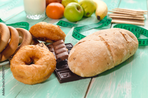 fruit with Cup of yogurt and bread separated by a measuring tape with chocolate and donuts on a green wooden background, concept of proper nutrition