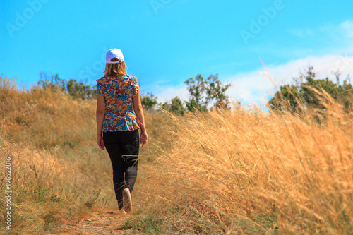 A girl in tights, a blouse and a white cap goes along the stony path among the bushes, grass and flowers against the blue sky.