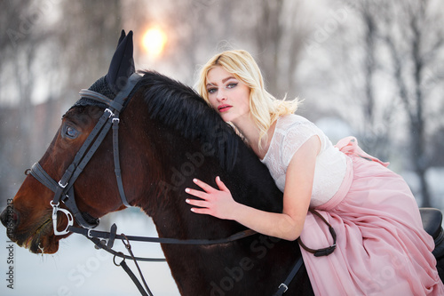 Young woman in rose dress riding her bay horse on winter field. Romantic or historical equestrian background with copy space